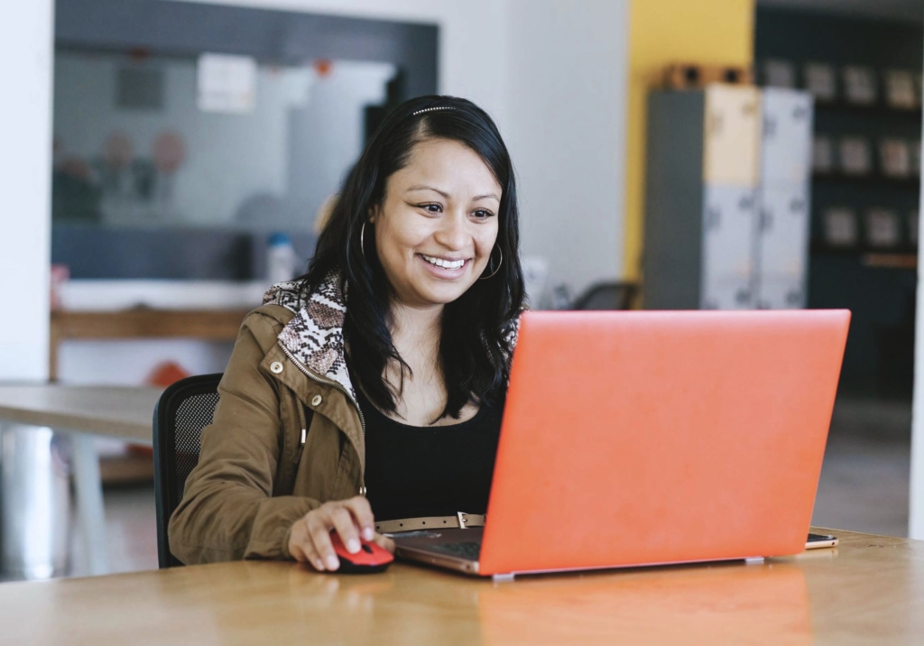 Photo of a woman smiling sitting at a table working on a laptop to showcase how technology can be adapted for accessibility services