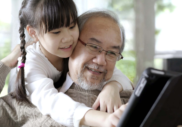 Photo of a man with a young girl hugging him over the shoulders as they look at a tablet while smiling