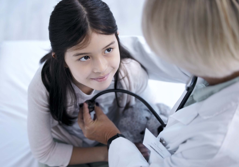 Photo of a child looking up at a woman doctor who is checking her heartrate with a stethoscope in a healthcare setting