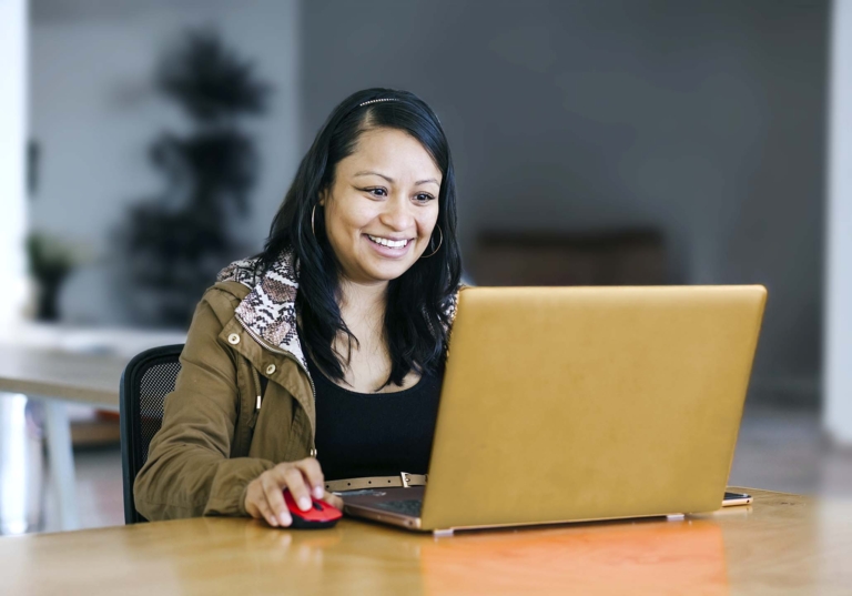 Photo of a woman smiling sitting at a table working on a laptop to showcase how technology can be adapted for accessibility services