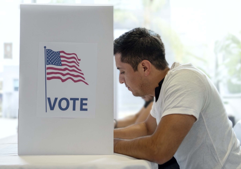 Photo of a man standing at an elections voting booth that has a graphic of a USA flag and reads "vote"