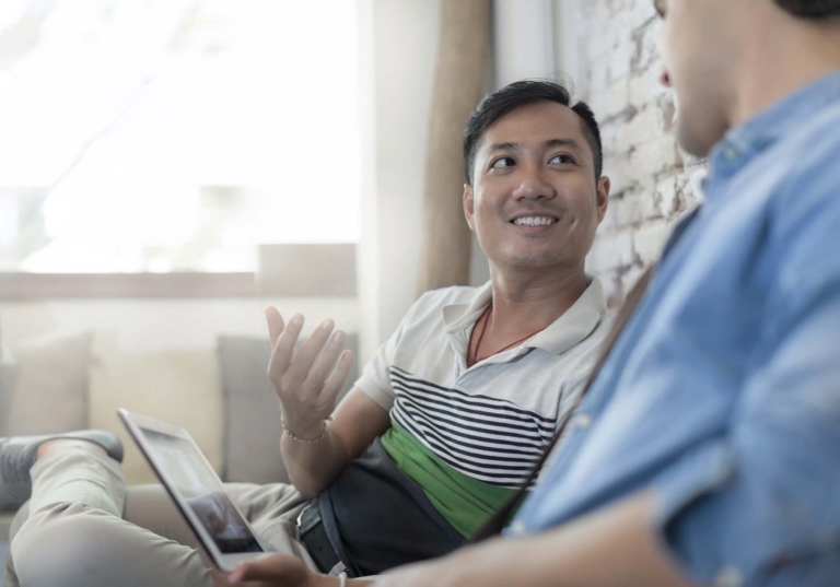 Photo of two men sitting together on a couch while looking at each other in conversation with a laptop